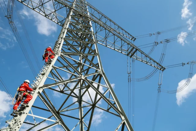 Zwei Monteure arbeiten an einem Hochspannungsmast beim Freileitungsbau vor blauem Himmel, Symbolbild zu den steigenden Strom-Umlagen 2027.