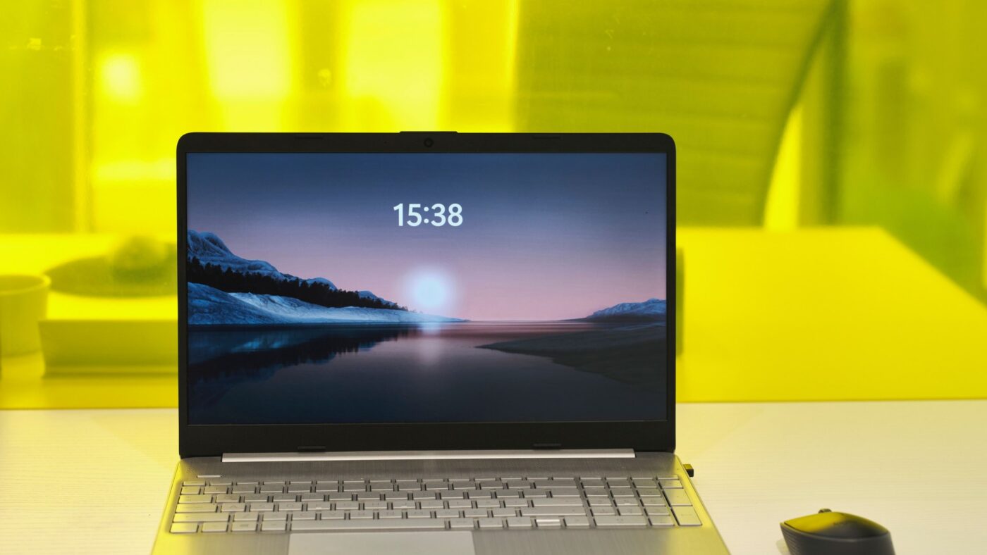 Silberfarbenes Notebook auf einem weißen Schreibtisch vor gelbem Hintergrund, auf dem Bildschirm eine Berglandschaft am See mit der Uhrzeit 15:38