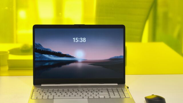 Silberfarbenes Notebook auf einem weißen Schreibtisch vor gelbem Hintergrund, auf dem Bildschirm eine Berglandschaft am See mit der Uhrzeit 15:38